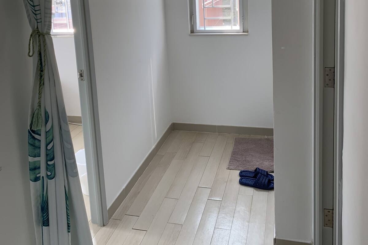 Una habitación con suelo de madera, una ventana con vistas a un edificio y una puerta que da a otra estancia.