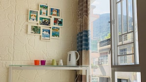 Une petite pièce avec un mur blanc, une fenêtre avec des rideaux, une table avec deux tabourets et une étagère avec des photos.