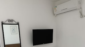 Une chambre à coucher comprenant un lit, un téléviseur fixé au mur, une table de chevet avec une télécommande, un climatiseur mural et un miroir avec un cadre.