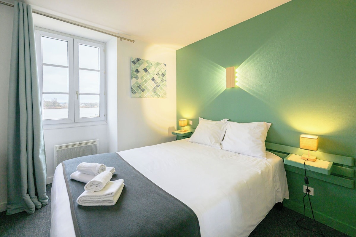 A hotel room with a bed, a nightstand with a lamp, a window with curtains, and a wall-mounted light fixture.