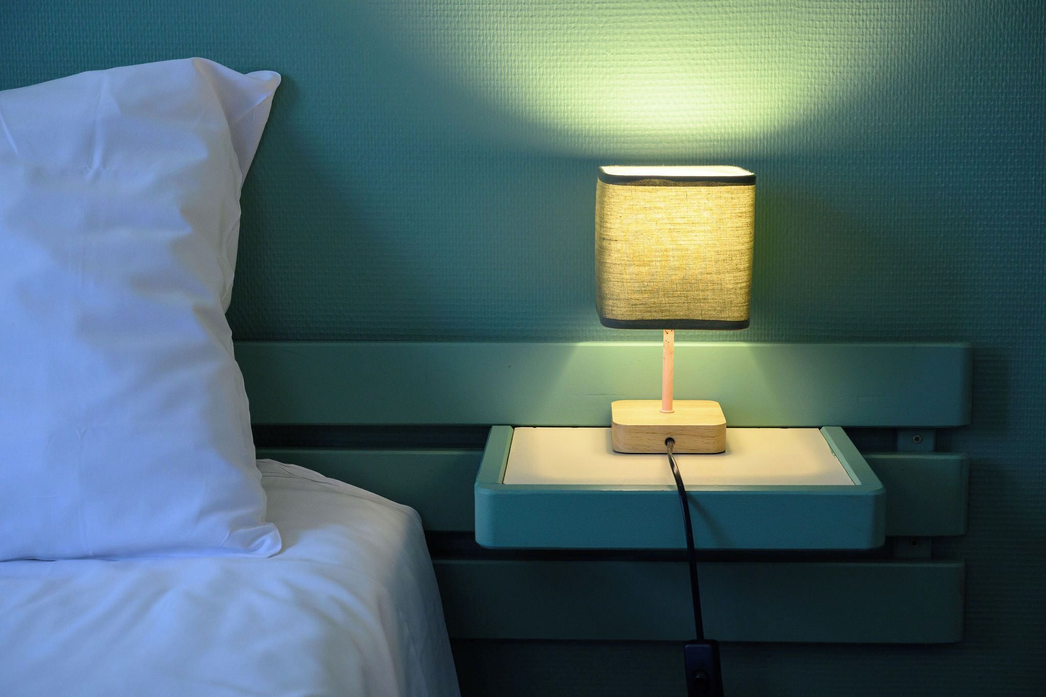 Una cama con sábanas y almohadas blancas, una mesita de noche con lámpara y una pared verde.