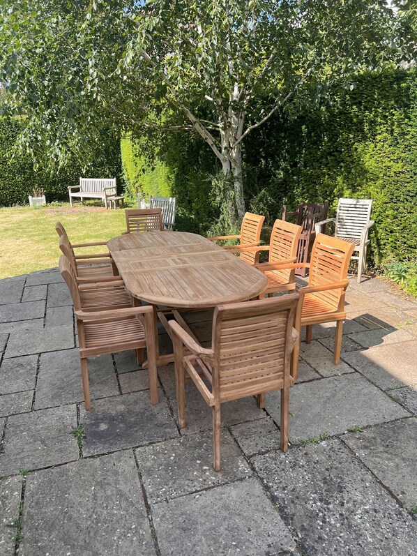 A wooden outdoor dining set with a table and six chairs, surrounded by a hedge and a bench in a garden setting.