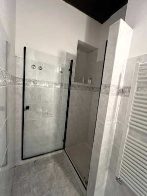 Un área de ducha con mampara de vidrio, paredes de azulejos y ducha en esquina.