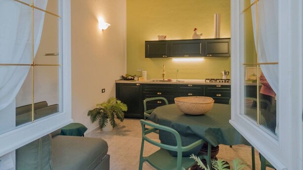 A compact kitchen with a green wall, a dining area with a table and chairs, and a window with curtains. - Picabon Apartment with AC Balcony (Monterosso al Mare)