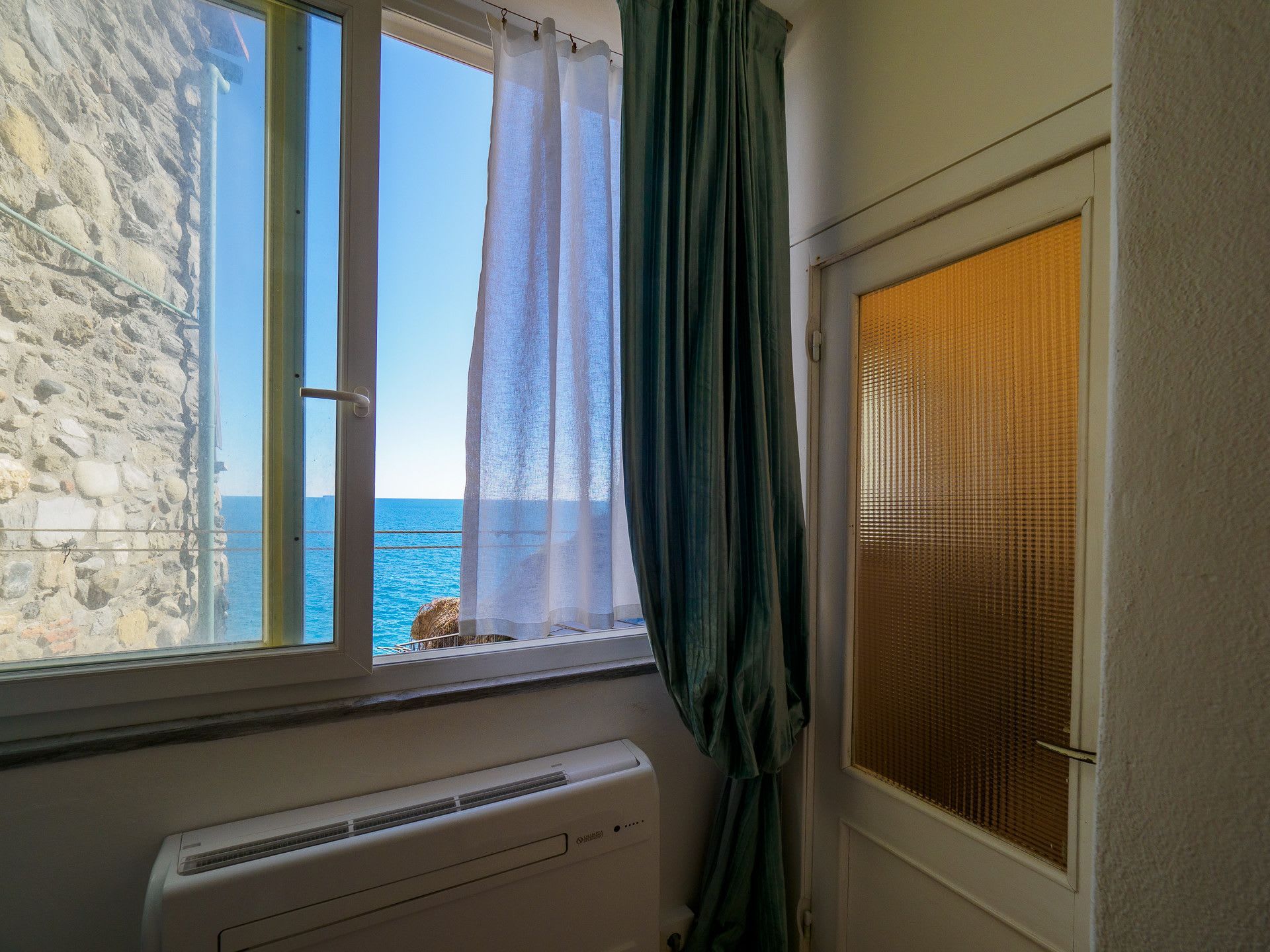 Una stanza con una finestra che si affaccia su una costa rocciosa, un termosifone e una porta con pannello decorativo.