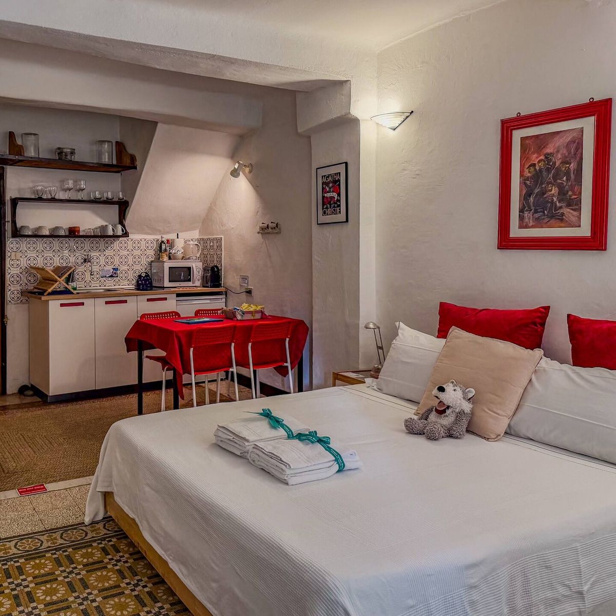 A hotel room with a bed, a desk with a red tablecloth, a microwave, and a small kitchenette.