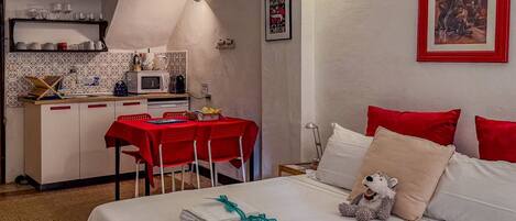 A hotel room with a bed, a desk with a red tablecloth, a microwave, and a small kitchenette.