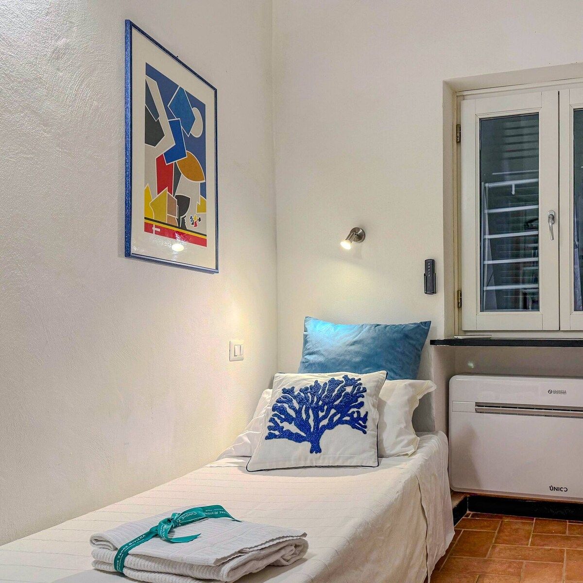 Un petit lit bien fait, avec un oreiller bleu, un mur blanc orné d’un tableau abstrait encadré, une fenêtre avec des stores et un appareil de climatisation blanc.