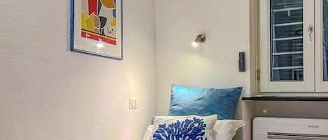 Un petit lit bien fait, avec un oreiller bleu, un mur blanc orné d’un tableau abstrait encadré, une fenêtre avec des stores et un appareil de climatisation blanc.