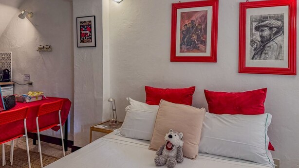 A bedroom with a bed, red and white pillows, a small stuffed animal, and artwork on the wall.