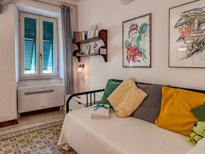 A bedroom with a bed, a window with shutters, a radiator, a shelf with books, and a wall with two framed artworks.