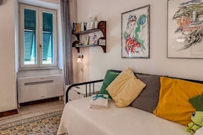 A bedroom with a bed, a window with shutters, a shelf with books and a framed painting, and a wall-mounted lamp.