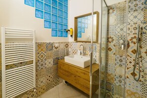 Casa de banho com lavatório branco, toucador de madeira, azulejos estampados, um porta-toalhas e cabine de duche em vidro.