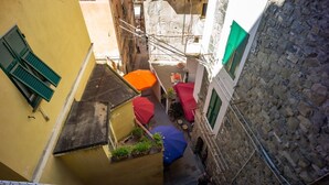 Un estrecho callejón con una pequeña zona de estar al aire libre, con sombrillas rojas y azules, un edificio amarillo con un persiano verde y un muro de piedra en un lateral.