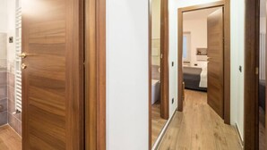 Um corredor com portas de madeira, soalho em azulejo e uma visão de um quarto com cama e mesa de cabeceira.