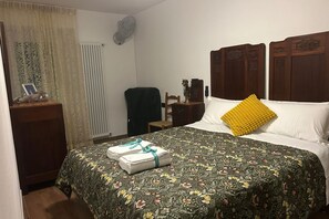 A hotel room with a bed, a chair, a desk with a picture frame, a radiator, and a fan.