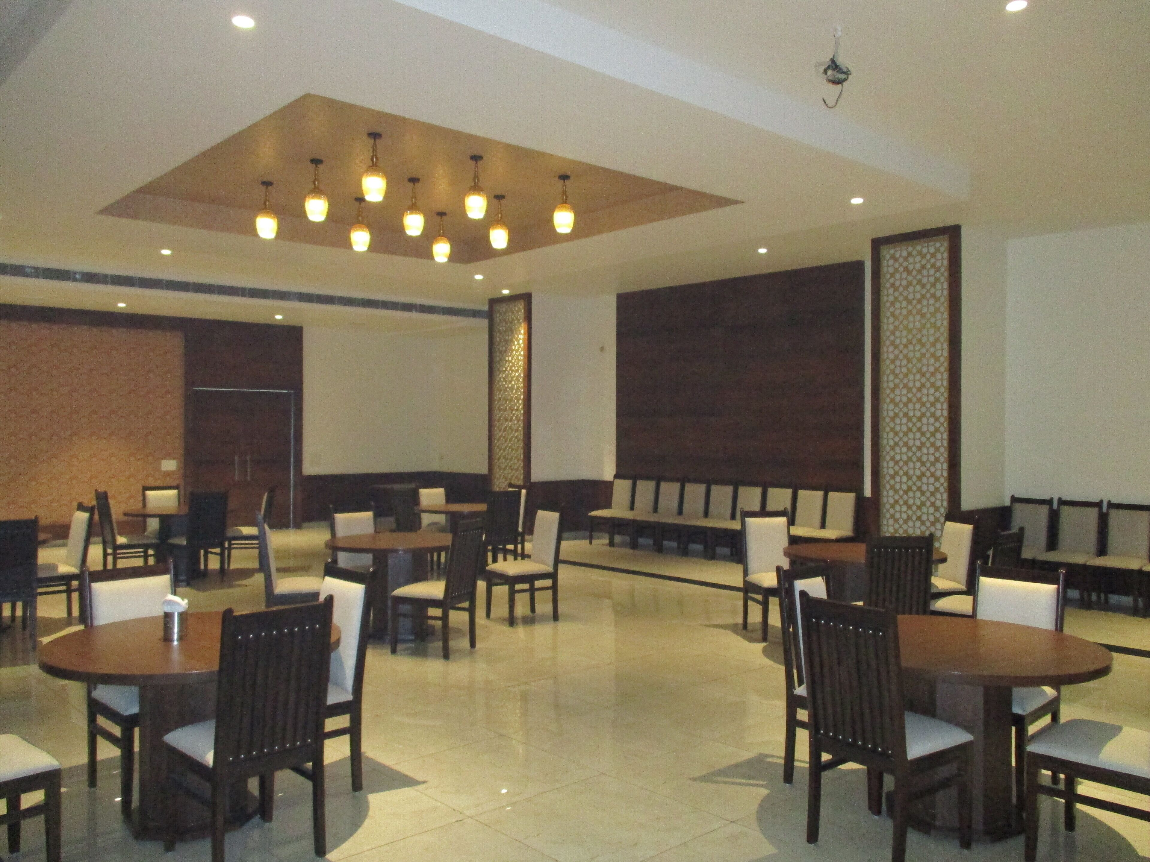 A spacious dining area with multiple round tables and chairs, a high ceiling with recessed lighting, and a large screen on one wall.