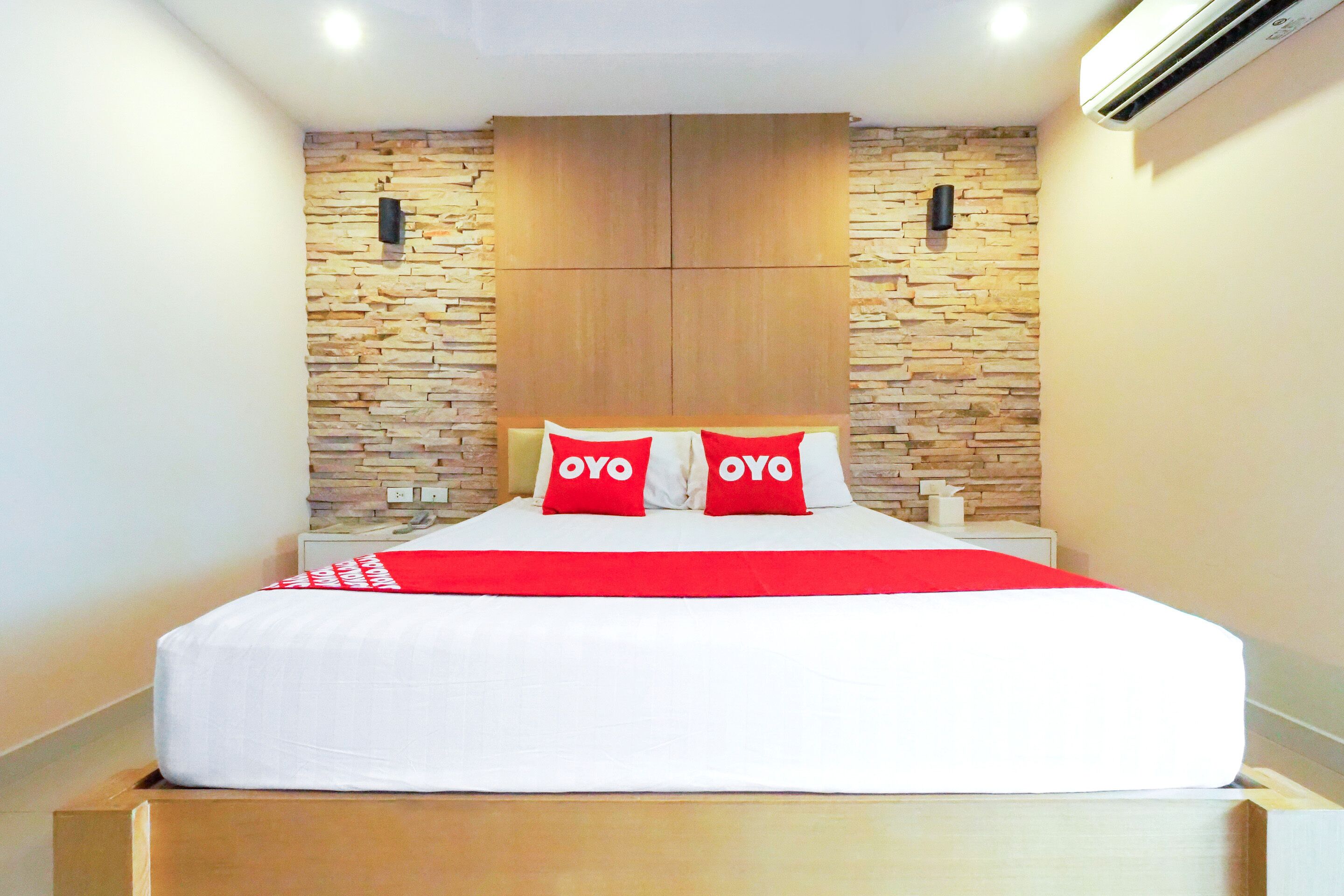 Una cama bien tendida con una colcha blanca y roja, dos almohadas con la palabra “OYO” en ellas y un cabecero de madera frente a una pared con diseño de piedra.