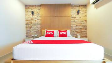 Una cama bien tendida con una colcha blanca y roja, dos almohadas con la palabra “OYO” en ellas y un cabecero de madera frente a una pared con diseño de piedra.