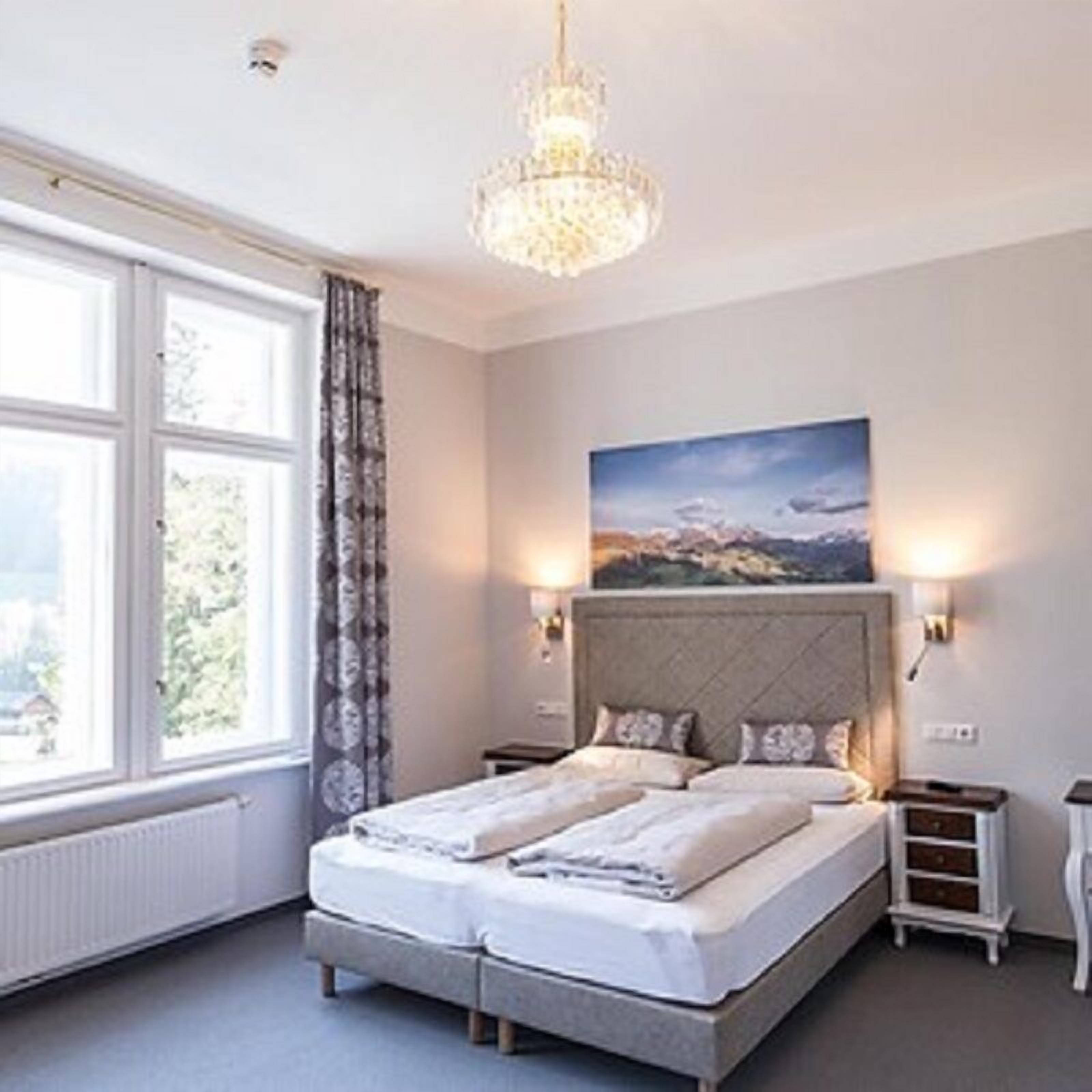 A bedroom with a large bed, a chandelier, a painting on the wall, and a window with curtains.