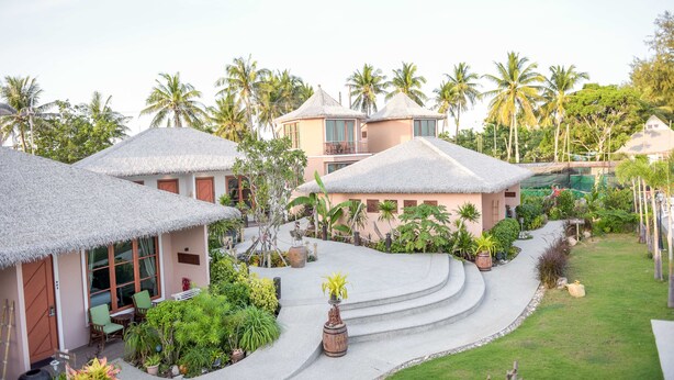 Um resort com chalés de telhado de palha, um edifício central com varanda e um jardim bem cuidado com palmeiras e diversas plantas.