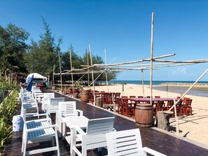 Un restaurant situé en bord de mer, avec des chaises longues blanches, des tables en bois, des parasols en bambou et une vue sur l’océan.