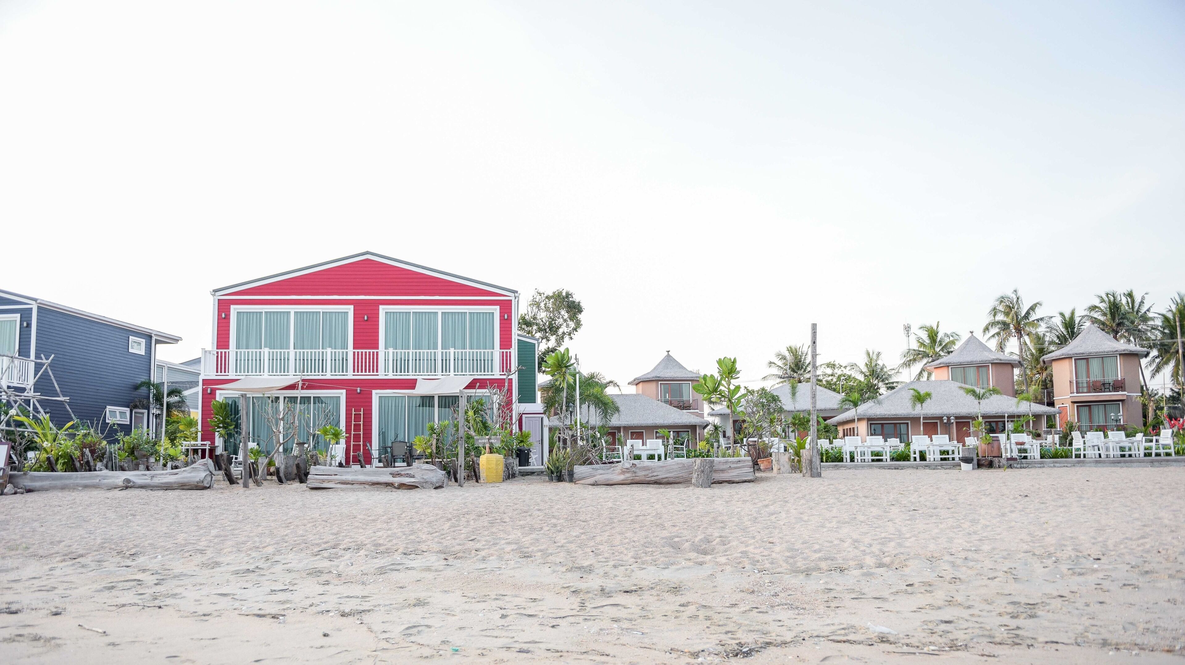 A beachfront property with a prominent red building featuring a balcony, surrounded by smaller structures and palm trees.