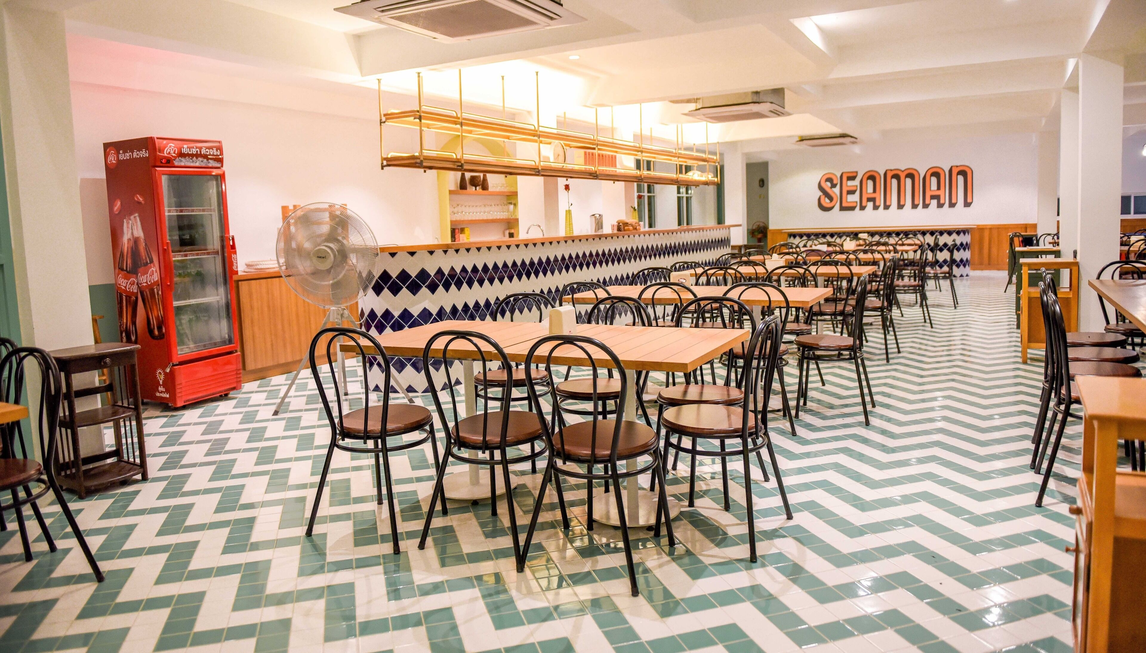 A cafeteria with multiple tables and chairs, a Coca-Cola refrigerator, and a wall with the word "SEAMAN".