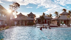Uma área de piscina com céu claro, palmeiras e edifícios com telhados de palha.