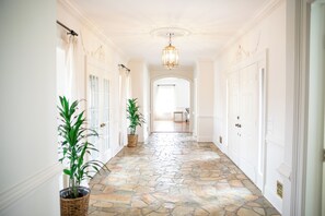 Ein Korridor mit Steinböden, weißen Wänden und einem Chandelier. Auf beiden Seiten befinden sich Blumentöpfe und Türen.