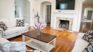 A living room with a white sofa, a coffee table, a fireplace, and a flat-screen TV mounted above it. - 12-Person House with Hot Tub on Spectacular Estate (Castleton)