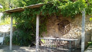 An outdoor patio with a stone wall, a pergola covered in greenery, a metal table with chairs, and a vintage wagon wheel decoration.