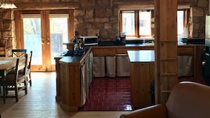L’intérieur d’un chalet rustique comprend une table à manger en bois, un canapé, un coin cuisine avec un mur en pierre et un sol en briques rouges.