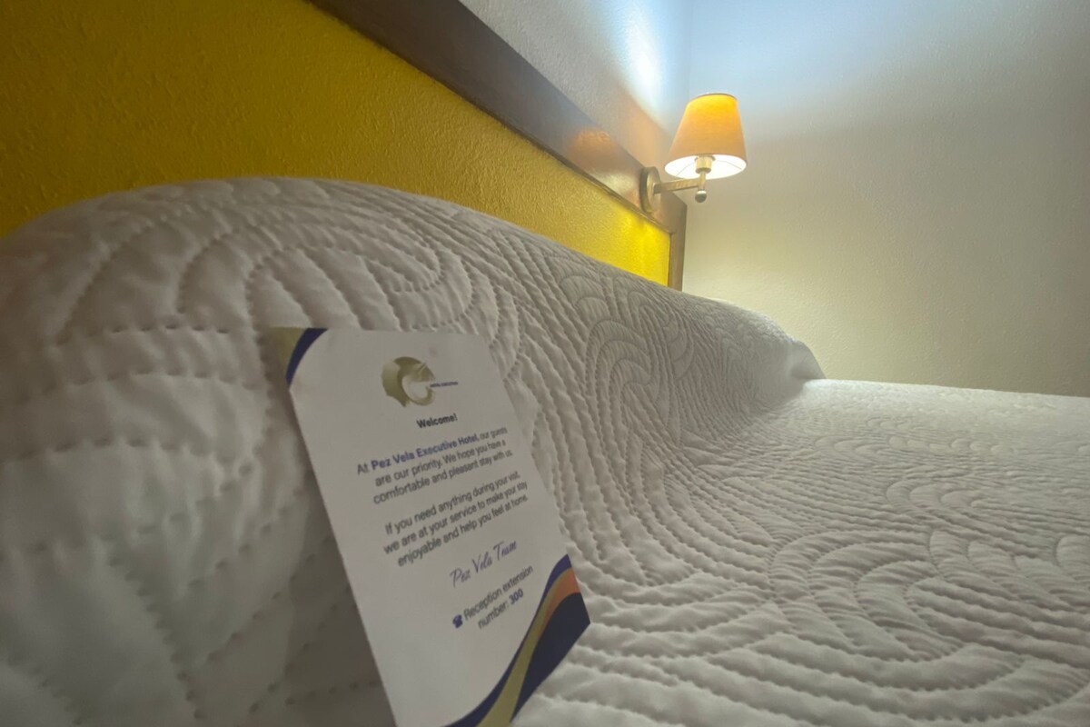 Habitación de hotel con una colcha blanca con textura, una almohada y una tarjeta de extensión de recepción.