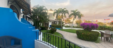A balcony with a blue wall, a chair, a railing, and a view of a garden with palm trees and purple flowers.