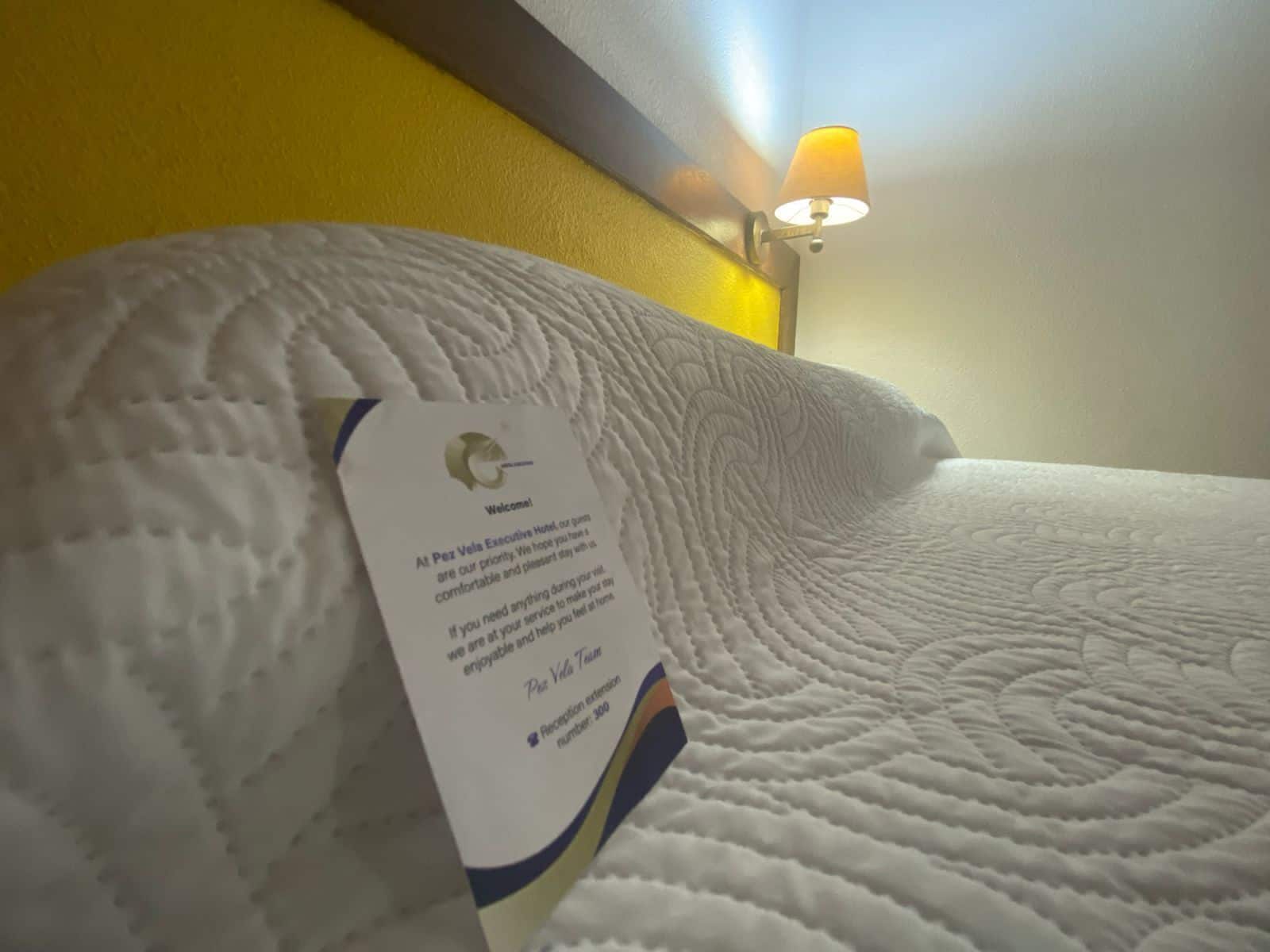 Une chambre d’hôtel avec un couvre-lit blanc à motifs, un oreiller et une carte de bienvenue posée sur le lit.
