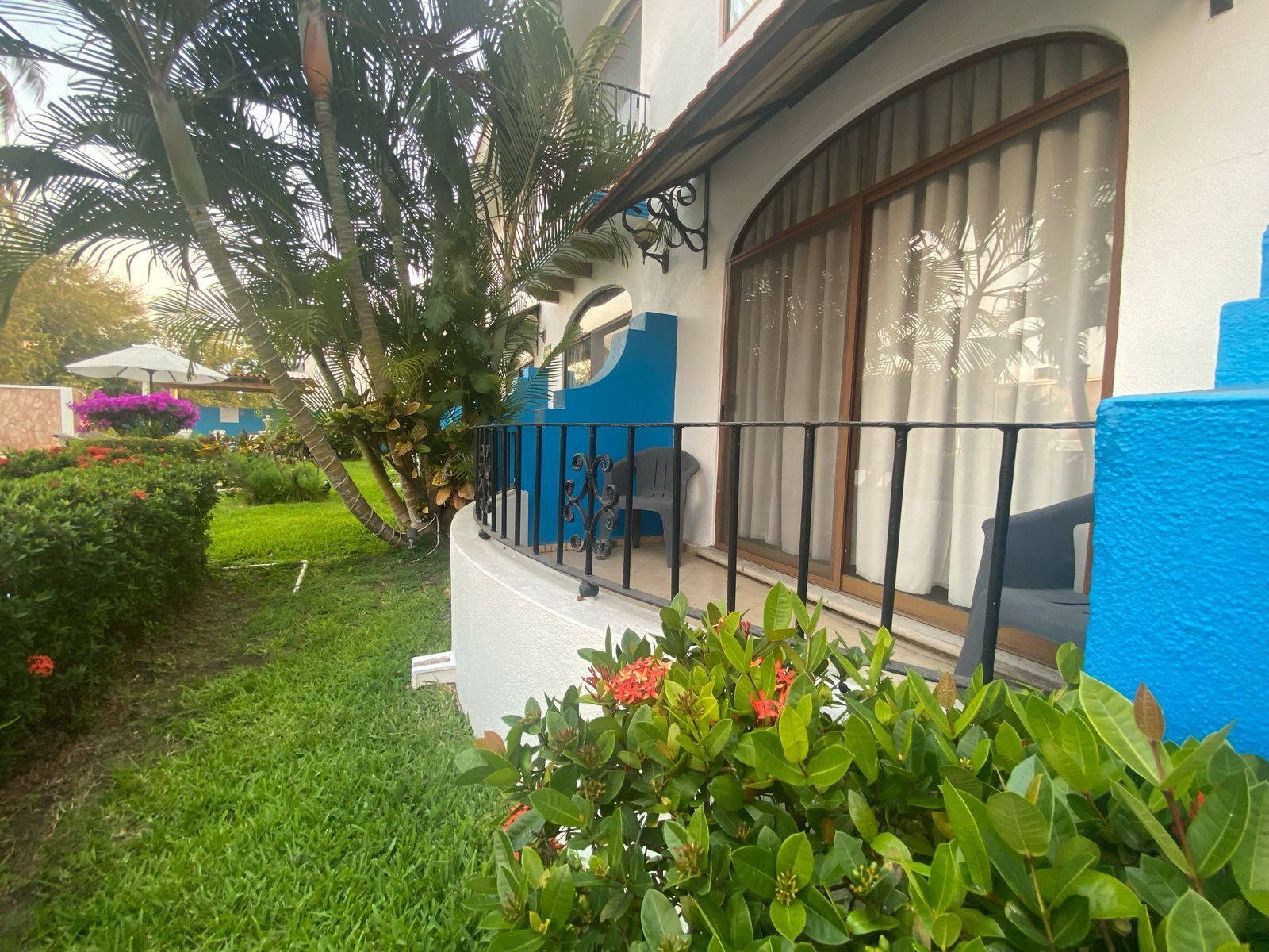 Una casa con fachada azul, un balcón con barandilla negra y un jardín con césped verde y flores rojas.