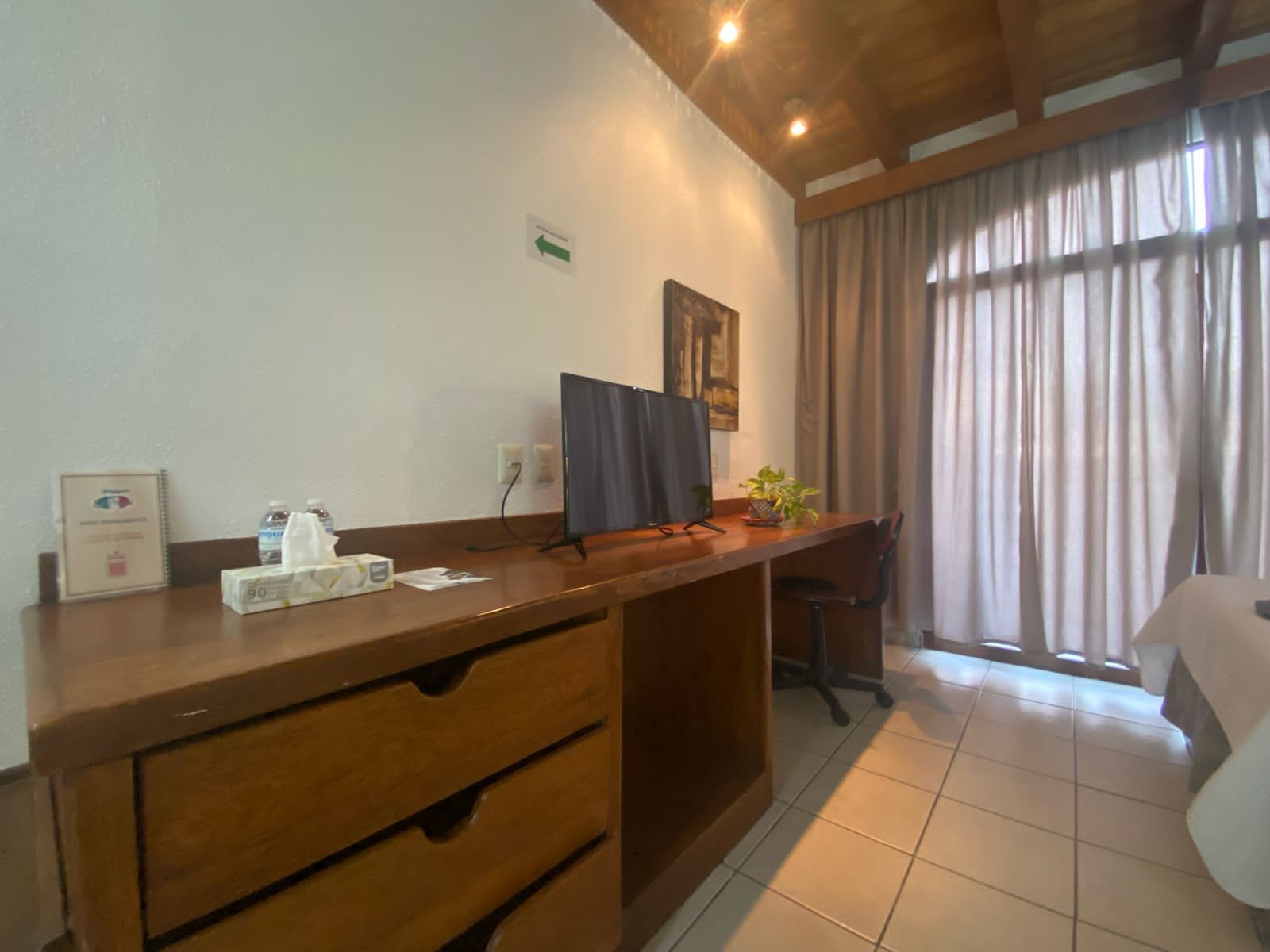 A hotel room with a wooden desk, a computer, a chair, a bed, and a window with curtains.