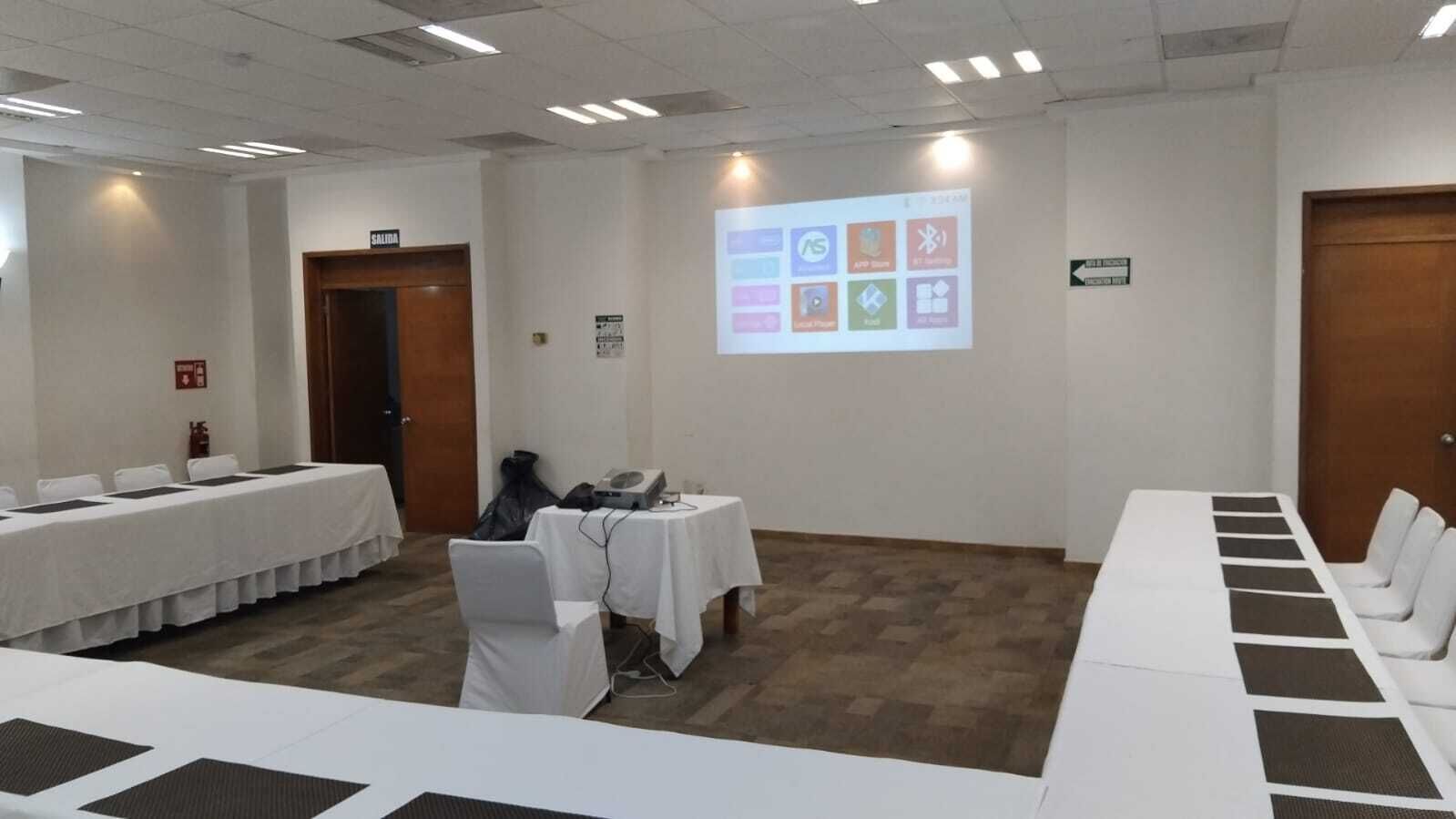 A conference room with a projector screen displaying icons, white tables with chairs, and a door labeled "SALIDA".