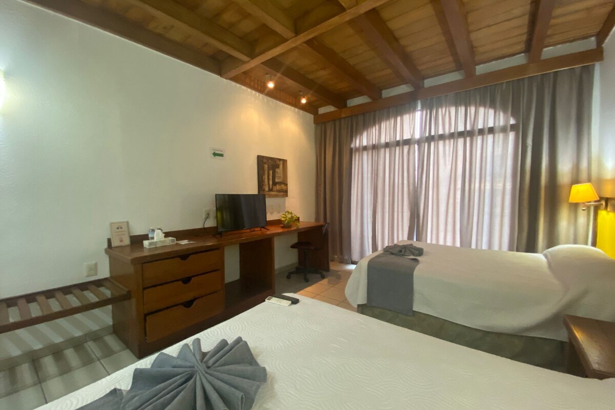 Habitación de hotel con cama, escritorio con computadora, silla, ventana con cortinas y lámpara.