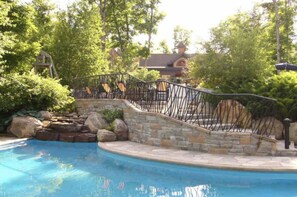 Ein Pool mit einer Veranda aus Stein und Metall, umgeben von üppiger Vegetation und Bäumen.