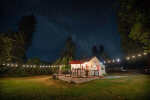 Uma casa branca com telhado vermelho e um deck, rodeada por árvores e luzes de Natal.