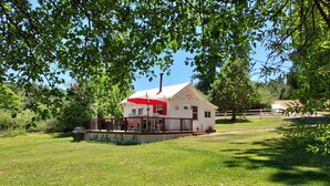 A small white cabin with a red roof and a deck, surrounded by lush green grass and trees.