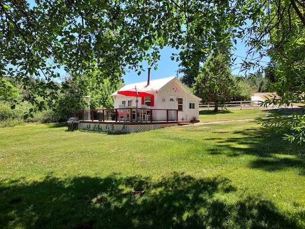 A small white cabin with a red roof and a deck, surrounded by lush green grass and trees.