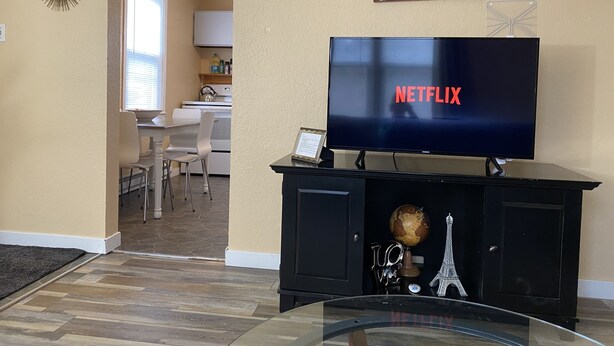 Una sala de estar con un televisor de pantalla plana mostrando 'NETFLIX', una mesa de centro de vidrio, un mueble de entretenimiento negro con objetos decorativos y una cocina visible a través de un vano abierto.