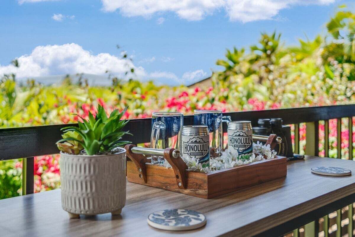 Una bandeja de madera con latas de cerveza Honolulu, una planta en maceta y un posavasos sobre la barandilla de un balcón.