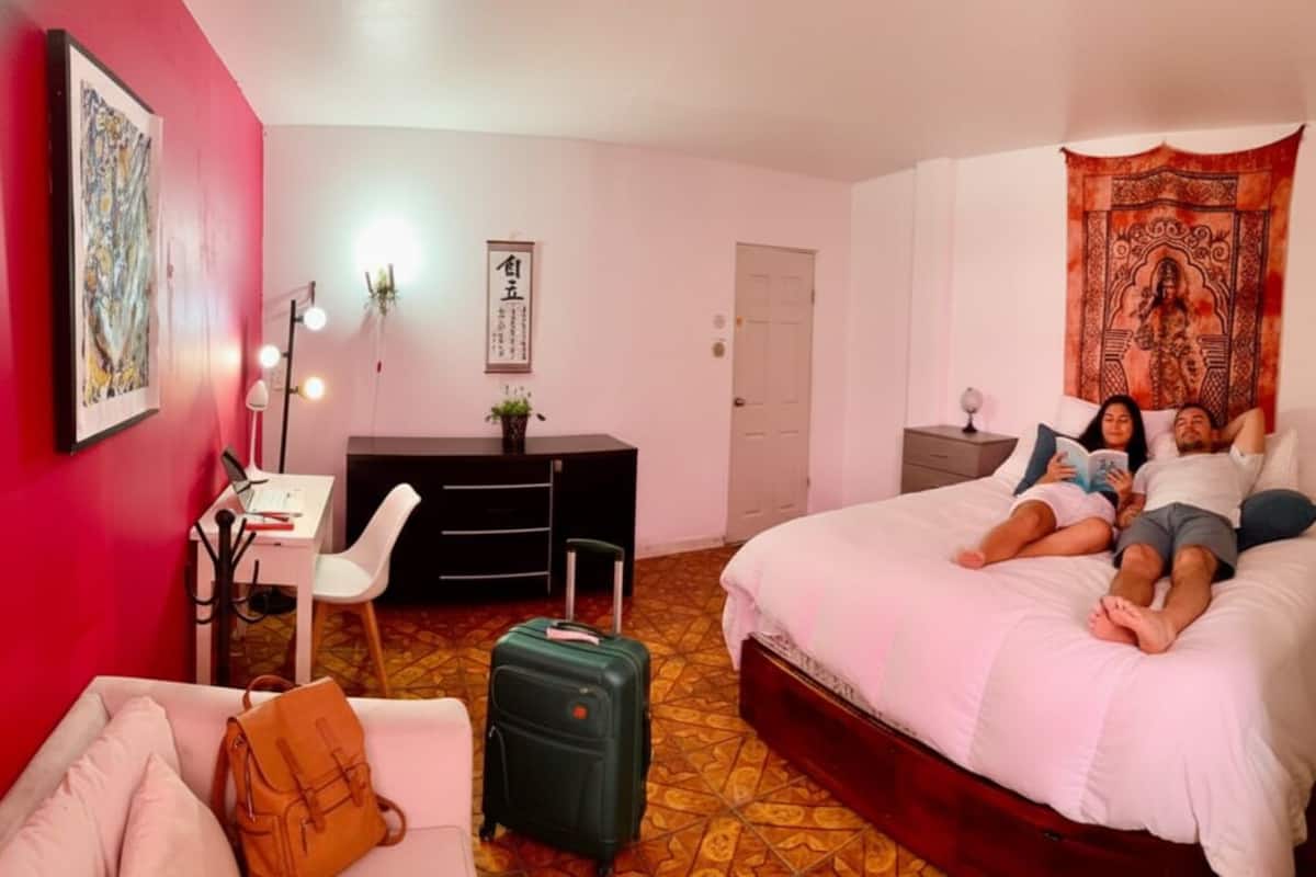 A hotel room with a bed, desk, chair, and a red wall with artwork.