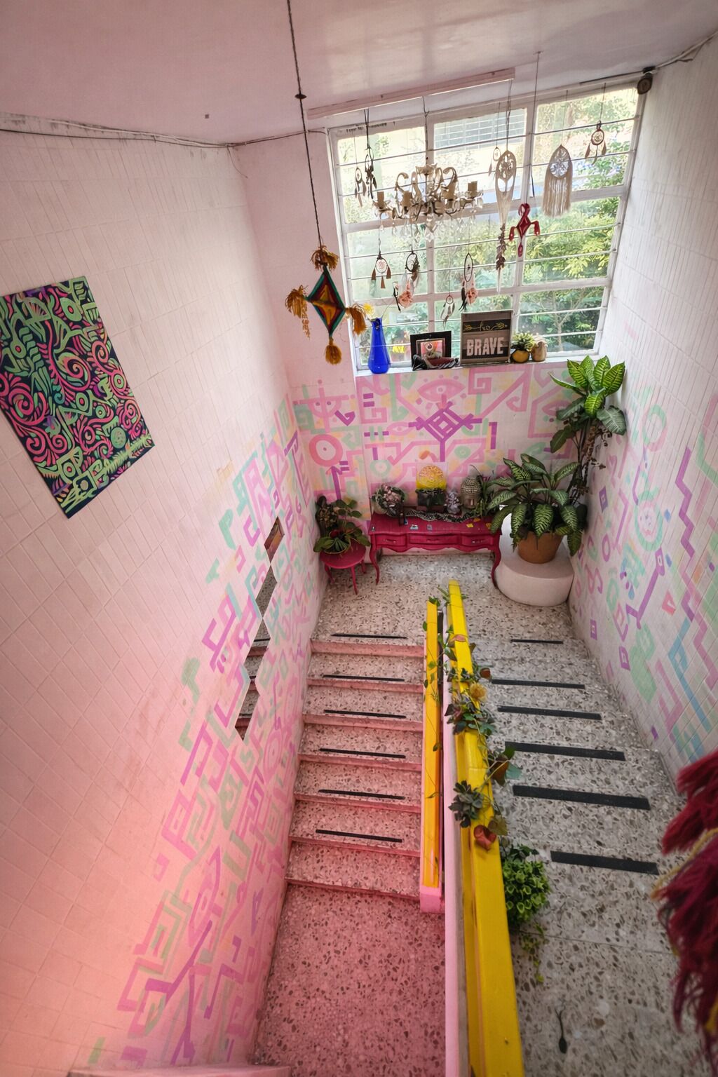 Una escalera con patrones geométricos coloridos en las paredes y un pequeño altar con plantas y un cartel que dice 'BRAVE'.