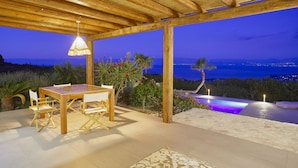 Eine Holzdielen-Terrasse mit Tisch und Stühlen, mit Blick auf einen Pool und eine schöne Aussicht auf Meer und Berge.