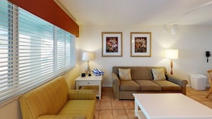 A cozy living room with a sofa, a small table, and two lamps. There are framed pictures on the wall and a window with blinds. - Holmes Beach | Couples Gulf Coast Retreat | Pool + Beach (Holmes Beach)